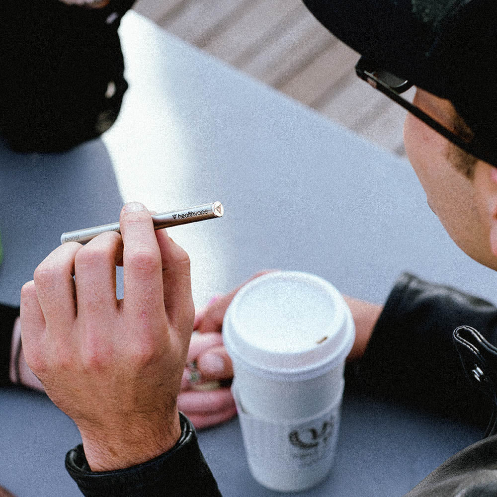 Man holding HealthVape Boost Berry Mint disposable vape pen with coffee cup at table