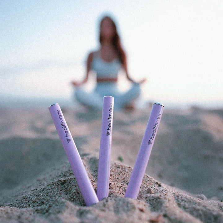 Three HealthVape Chill Lavender disposable vape pens standing in sand with blurred yoga figure