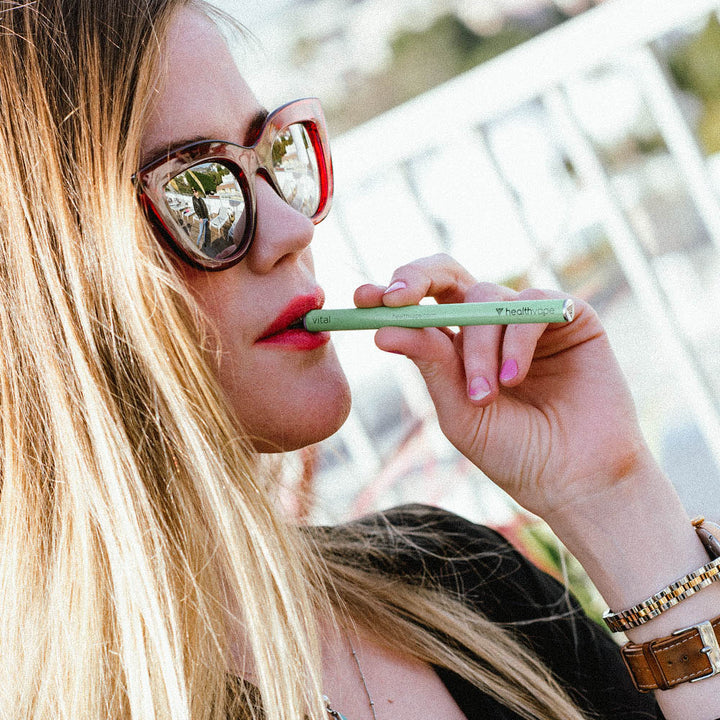 Woman wearing sunglasses outdoors using a green HealthVape VITAL disposable inhaler.